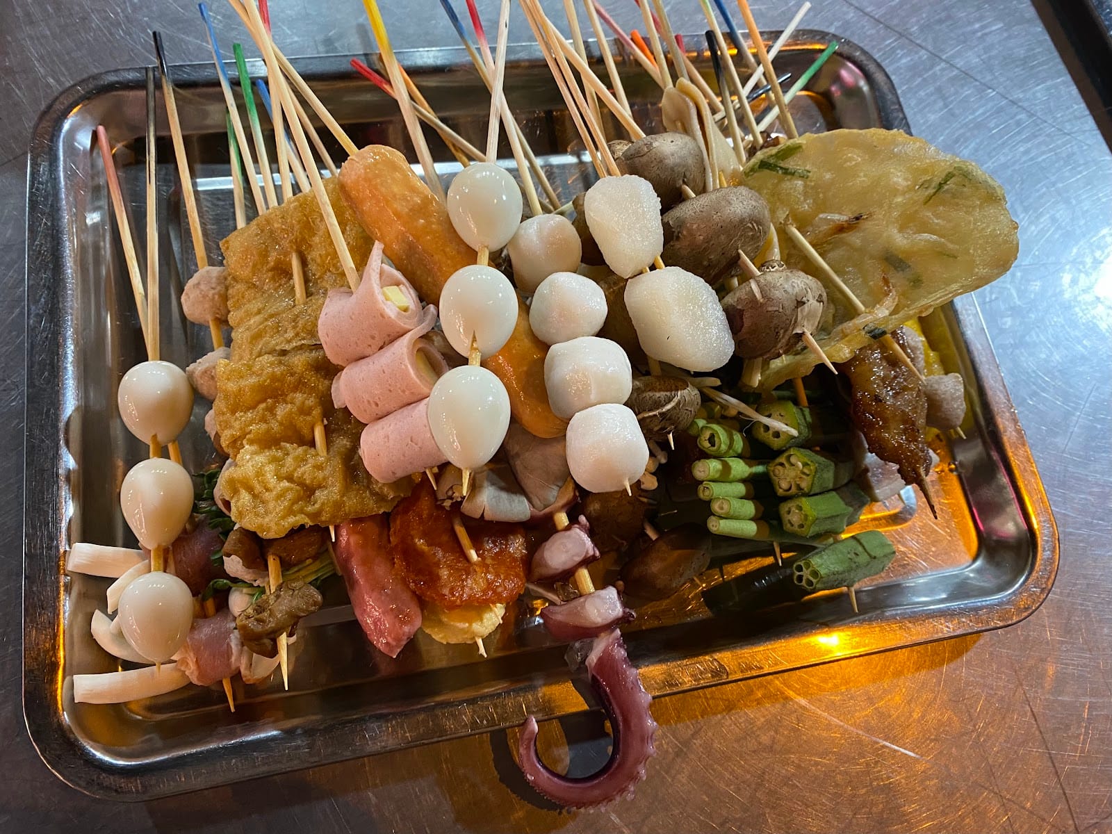 A steel tray piled with picked skewers: quail eggs, fish balls, sausages, mushrooms, okra and octopus