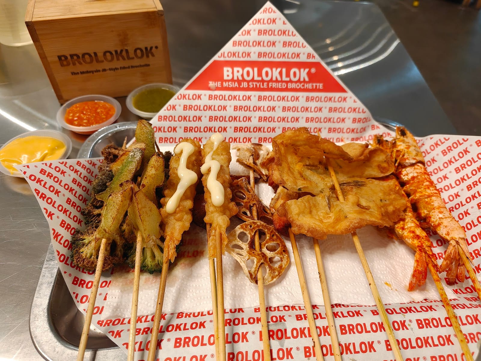 A tray of fried skewers on BROLOKLOK-printed paper — broccoli, baby corn, lotus root, fish cake and prawns — with three dips and a BROLOKLOK wooden box
