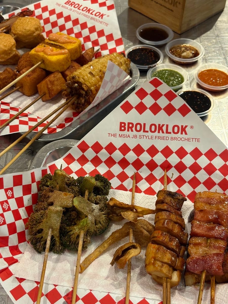 Fried skewers — broccoli, mushroom, fish cake, bacon rolls and grilled corn — on red-and-white BROLOKLOK paper, with six dipping sauces behind