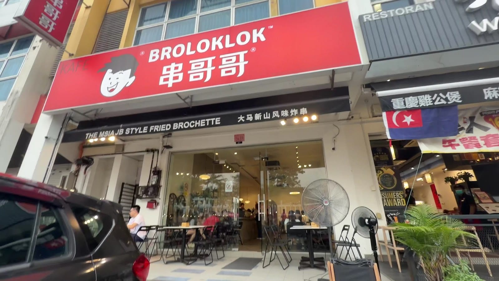 The Broloklok 串哥哥 shopfront on Jalan Mutiara Emas 2A: a red signboard with the cartoon mascot, the banner “The MSIA JB Style Fried Brochette” and tables out front
